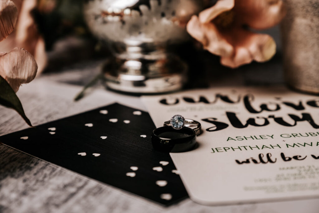 wedding ring that is blue stone and black wedding band on top of white black and gold invitation with pink flowers in background