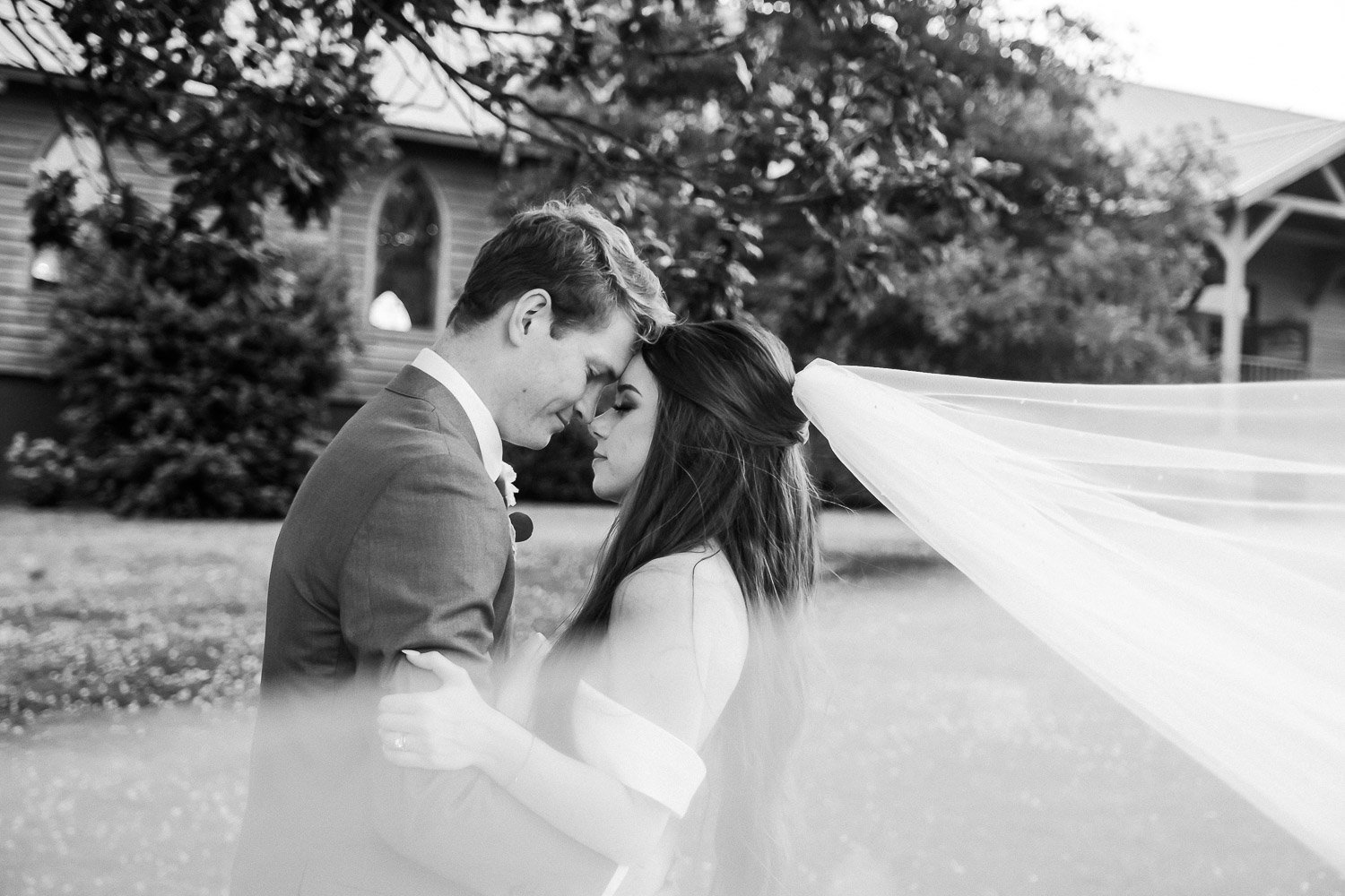 A bride and groom facing each other embracing as husband and wife with their heads touching and the brides veil swooping in front of the camera lens.