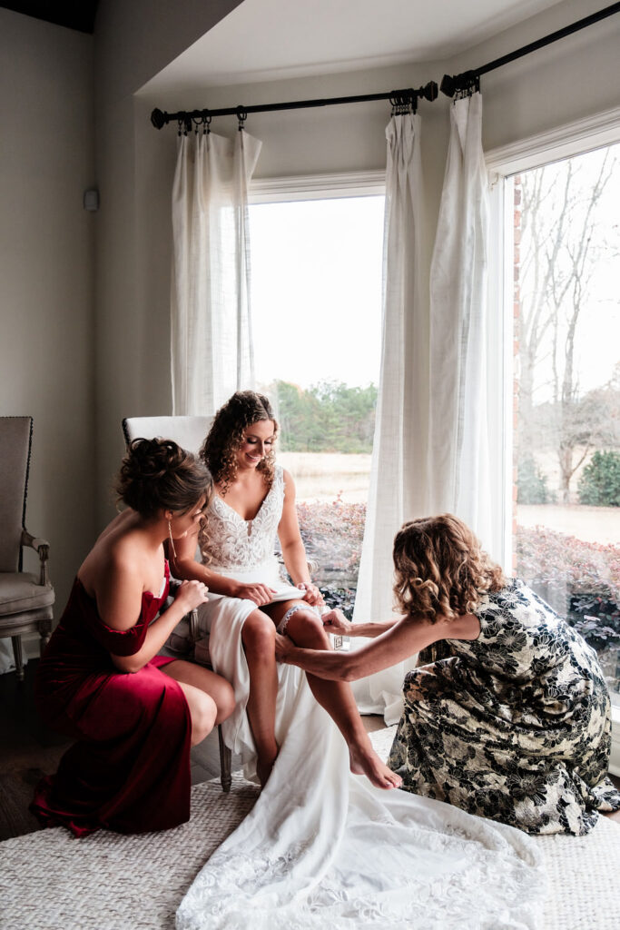 Bride getting ready with her mom and sister helping put on shoes and accessories before her wedding day.